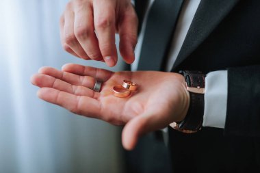 Bir adam elinde iki alyans tutuyor. Yüzükler altın ve yüksek kalitede görünüyor. Adam takım elbise giymiş ve kravat takmış. Resmi bir etkinliğe katıldığı izlenimini veriyor.