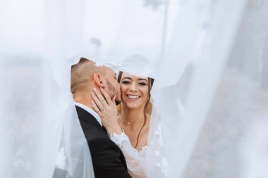 Gelin ve damat beyaz bir duvağın önünde öpüşüyorlar.