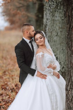 Bir gelin ve damat bir ağacın önünde sarılıyorlar. Gelin beyaz bir elbise giyiyor ve damat da takım elbise giyiyor.