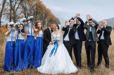 Bir grup insan, ortasında bir gelin ve damat olan bir resim için poz veriyor. Gelin mavi bir elbise giyiyor ve damat bir takım elbise giyiyor. Grup gülümsüyor ve birbirine sarılıyor.