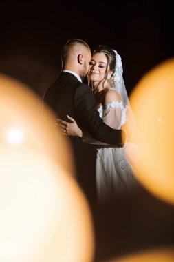 Bir gelin ve damat düğün fotoğrafında birbirlerine sarılıyorlar. Gelin beyaz bir elbise giyiyor ve damat bir takım elbise giyiyor. Fotoğrafın sıcak ve romantik bir havası var, aşkı yakalıyor.