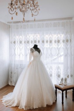 Üzerinde avize olan bir odada beyaz bir gelinlik sergileniyor. Elbise uzun ve dalgalı, ve bir sandalyede oturuyor. Odanın sıcak ve davetkar bir atmosferi var.