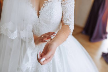 Bir gelin düğünü için hazırlanıyor ve gelinliğini giyiyor. Dantel peçeli beyaz bir elbise giyiyor ve elinde bir bilezik tutuyor.