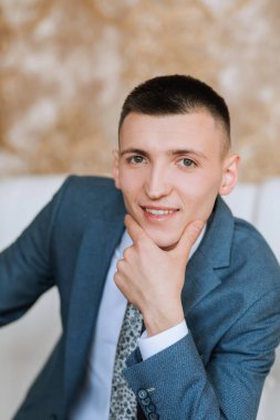 Mavi takım elbiseli ve kravatlı bir adam kanepede oturuyor ve kameraya bakıyor. Yüzünde ciddi bir ifade var ve elini çenesine dayıyor. Profesyonellik ve resmiyet kavramı