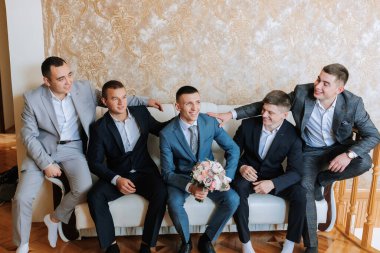 Bir grup adam kanepede oturuyor, takım elbiseli ve kravatlı. Adamlardan biri bir buket çiçek tutuyor. Manzara rahat ve dostça, çünkü erkekler birbirlerinin arkadaşlığından hoşlanıyorlar.