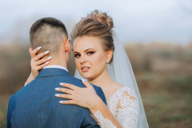 Bir gelin ve damat bir tarlada birbirlerini kucaklıyor. Gelin beyaz bir peçe takıyor ve damat mavi bir takım giyiyor.