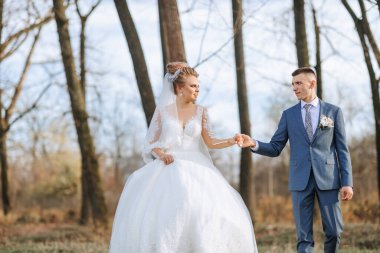 Bir gelin ve damat el ele bir ormanda yürüyorlar. Gelin beyaz bir elbise giyiyor ve damat mavi bir takım elbise giyiyor. Sahne romantik ve huzurlu.