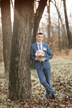 Takım elbiseli bir adam elinde bir buket çiçek olan bir ağacın yanında duruyor. Sahne sakin ve huzur dolu. Uzağa bakan adamla birlikte.