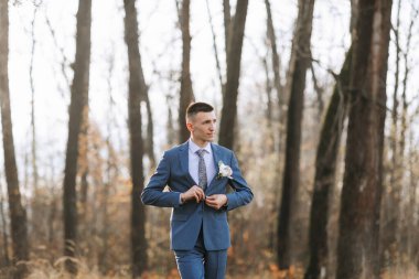 Mavi takım elbiseli bir adam ormanda yürüyor. O bir kravat takıyor ve bir çiçek tutuyor. Sahne sakin ve huzurlu, adam ileriye bakıyor ve orman doğal bir zemin sağlıyor.