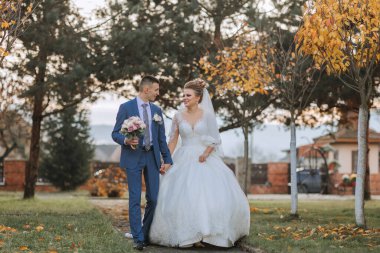 Bir gelin ve damat parkta bir patikada yürüyorlar. Gelin beyaz bir elbise giyiyor ve damat mavi bir takım elbise giyiyor. El ele tutuşuyorlar ve mutlu ve aşık görünüyorlar.