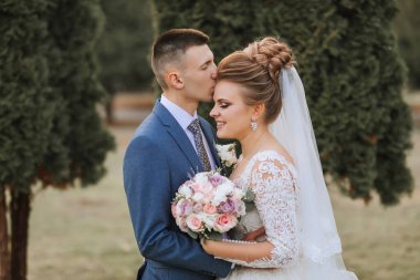 Bir gelin ve damat birbirlerine sarılıp öpüşüyorlar. Gelin elinde bir buket çiçek tutuyor.