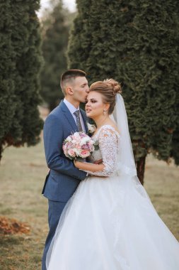 Gelin ve damat bir tarlada duruyorlar, gelin beyaz bir elbise giyiyor ve damat mavi bir takım elbise giyiyor.