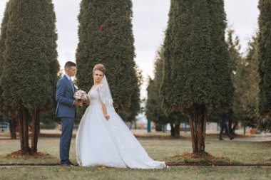 Bir gelin ve damat bir sıra ağacın önünde duruyor. Gelin beyaz bir elbise giyiyor ve damat mavi bir takım elbise giyiyor. Ellerinde bir buket ve küçük bir buket tutuyorlar.