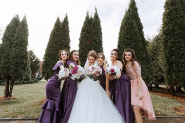 Bir grup kadın bir ağacın önünde fotoğraf için poz veriyor. Hepsi mor elbiseler giyiyor ve ellerinde çiçekler var. Sahne neşeli ve kutlayıcıdır.