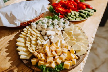 Çeşitli peynir ve sebzelerle dolu bir masa. Peynirler küçük parçalara ayrılır ve ahşap bir tabakta dizilir. Sebzeler arasında salatalık, domates ve biber var.