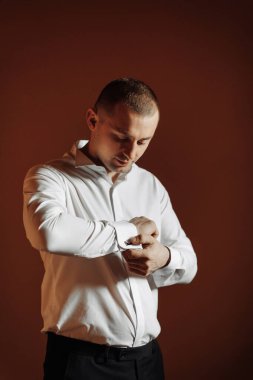 Beyaz gömlekli bir adam gömleğini düzeltiyor. Adam siyah bir takım elbise giyiyor ve kırmızı bir duvarın önünde duruyor.