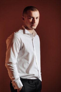 Beyaz gömlekli ve siyah pantolonlu bir adam kırmızı arka planın önünde duruyor. Ciddi ve odaklanmış görünüyor.