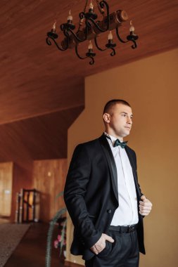 Siyah takım elbiseli ve beyaz gömlekli bir adam avizenin önünde duruyor. Papyon takıyor ve resmi bir etkinliğe hazırlanıyor.