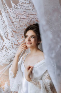 Beyaz bir perdenin önünde beyaz bir elbise ve beyaz bir peçe takan bir kadın var. O, yüzünde bir gülümsemeyle kameraya bakıyor. Mutluluk ve kutlama kavramı