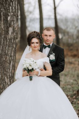 Bir gelin ve damat ormanda fotoğraf çektirmek için poz veriyorlar. Gelin bir buket çiçek tutuyor ve damat yeşil bir kravat takıyor. Sahne romantik ve samimi.