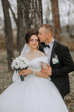 Bir gelin ve damat ormanda sarılıyor. Gelin elinde bir buket çiçek tutuyor.