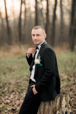 Siyah takım elbiseli ve beyaz gömlekli bir adam ormanda fotoğraf çektirmek için poz veriyor. Yakasına yeşil bir çiçek ve papyon takıyor. Sahne zarif ve romantik.