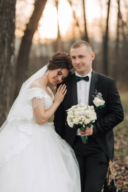 Bir gelin ve damat ormanda fotoğraf çektirmek için poz veriyorlar. Gelin beyaz bir elbise giyiyor ve damat siyah bir takım elbise giyiyor. Bir buket çiçek ve çiçek tutuyorlar.