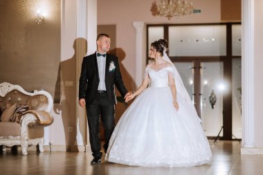 Bir gelin ve damat bir düğün töreniyle koridorda yürüyorlar. Gelin beyaz bir elbise giyiyor ve damat da takım elbise giyiyor.