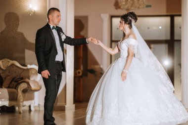 Bir gelin ve damat bir balo salonunda dans ediyorlar. Gelin beyaz bir elbise giyiyor ve damat bir takım elbise giyiyor. Sahne mutlu ve kutlama dolu.