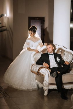 Bir gelin ve damat kanepede oturuyor. Gelin beyaz bir elbise giyiyor ve damat siyah bir takım giyiyor.