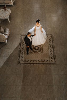 Bir gelin ve damat bir binanın önündeki halının üzerinde duruyor. Gelin beyaz bir elbise giyiyor ve damat siyah bir takım giyiyor.