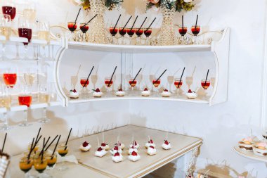 Şarap bardakları ve fincanlar dahil çeşitli tatlı ve içeceklerle dolu beyaz bir masa. Masa, tatlı ve içeceklere odaklanarak bir parti ya da toplanma için hazırlanmıştır.