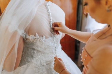 Bir kadın düğünü için hazırlanıyor ve annesi elbisesine yardım ediyor. Gelin, dantel yakalı ve peçeli beyaz bir elbise giyiyor. Anne gelinin elbisesini tutuyor.