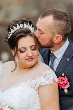 Bir gelin ve damat birbirlerinin yanaklarını öpüyor. Gelin taç takıyor ve damat da takım elbise giyiyor.