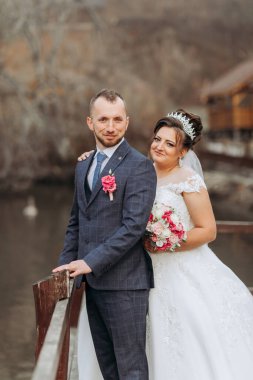 Bir adam ve bir kadın rıhtımda fotoğraf çektirmek için poz veriyorlar. Kadın beyaz bir elbise giyerken adam takım elbise ve kravat takıyor. İkisi de gülümsüyor ve birlikte olmaktan mutlu görünüyorlar.