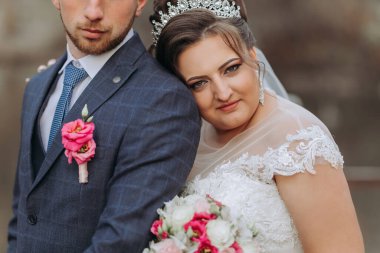 Bir gelin ve damat fotoğraf için poz veriyor. Gelin beyaz bir elbise ve taç giyiyor. Damat mavi bir takım elbise ve yakasında pembe bir çiçek giyiyor. Sahne mutlu ve romantik.