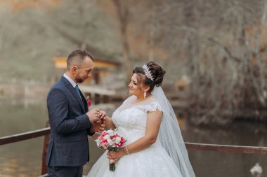 Bir gelin ve damat rıhtımda duruyorlar, alyanslarını takıyorlar. Gelin beyaz bir elbise giyiyor ve damat mavi bir takım elbise giyiyor. Gelin elinde bir buket çiçek tutuyor.