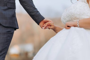 Bir gelin ve damat düğün fotoğrafında el ele tutuşuyorlar. Gelin beyaz bir elbise giyiyor ve damat bir takım elbise giyiyor. Fotoğraf, çift arasında bir sevgi ve bağlılık anı yakalıyor.