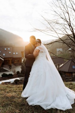 Bir gelin ve damat çimenli bir yamaçta duruyorlar, üzerlerinde güneş parlıyor.