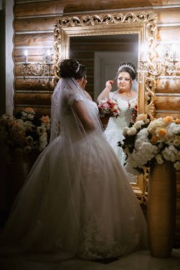 Bir gelin aynanın önünde duruyor, düğününe hazırlanıyor. Beyaz bir elbise ve taç giyiyor.