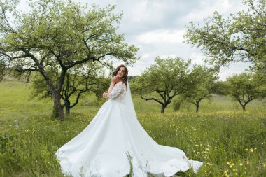 Beyaz elbiseli bir kadın çimlerin üzerinde duruyor. Alan ağaçlarla çevrili ve gökyüzü bulutlu. Kadın bir fotoğraf için poz veriyor ve görüntünün genel havası sakin.