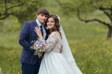 Bir gelin ve damat tarlada fotoğraf çektirmek için poz veriyorlar. Gelin beyaz bir elbise giyiyor ve damat mavi bir takım elbise giyiyor. Çiçek ve buket tutuyorlar. Sahne mutlu ve romantik.