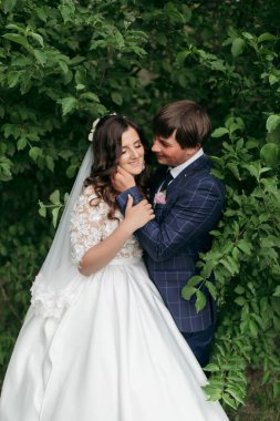 Bir gelin ve damat ormanda sarılıyor. Gelin beyaz bir elbise giyiyor ve damat mavi bir takım giyiyor.