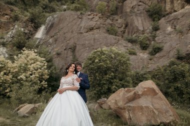 Bir gelin ve damat büyük bir kayanın önünde duruyor. Gelin beyaz bir elbise giyiyor ve damat mavi bir takım elbise giyiyor. İkisi de gülümsüyor ve kameraya bakıyor.