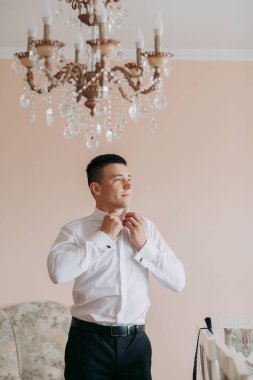 Beyaz gömlekli bir adam avizenin önünde giyiniyor. Sahne zarif ve sofistike.