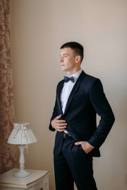 Takım elbiseli ve kravatlı bir adam lambanın önünde duruyor. Papyon takıyor ve resmi bir etkinliğe hazırlanıyor.