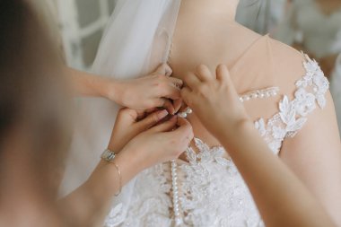 Bir kadın elbisesini başka iki kadına yaptırıyor. Gelin dantelli ve incili beyaz bir elbise giyiyor.