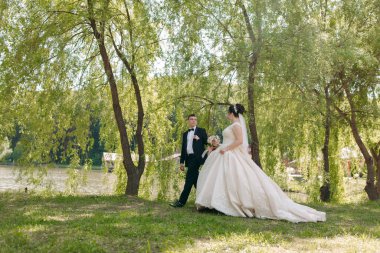Bir gelin ve damat, ağaçlar ve bir su kütlesi ile çevrili bir parkta yürüyorlar. Gelin uzun, beyaz bir elbise giyiyor ve damat siyah bir takım giyiyor. Huzur dolu bir sahne.