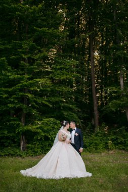 Gelin ve damat ormanda duruyorlar, gelin beyaz bir elbise giyiyor ve damat takım elbise giyiyor.