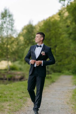 Siyah takım elbiseli ve beyaz gömlekli bir adam yolda yürüyor. Yakasına pembe bir çiçek takmış.